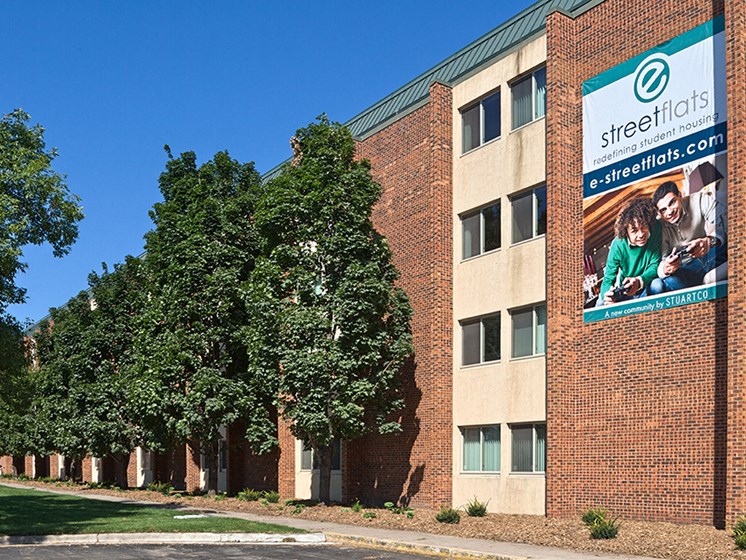 outdoor view of property with tan and brown bricks, tall green trees, and a sign on the building that says 